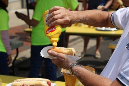 A person squeezes mustard onto a hot dog at an outdoor event, with others in the background.
