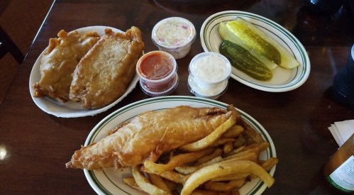 A plate of fried fish with fries, two pieces of fried fish, pickles, and dipping sauces on a wooden table.