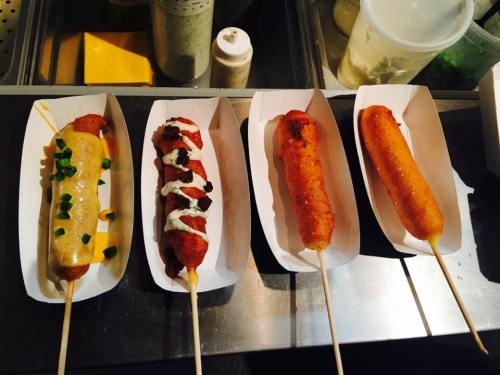 Four different corn dogs on paper trays, topped with various sauces and garnishes, displayed on a counter.
