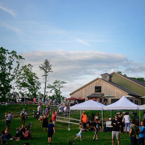 A lively outdoor event at a farm with tents, people mingling, and a rustic building under a blue sky.