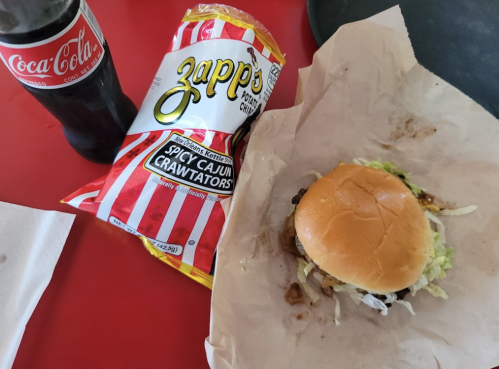 A burger with lettuce on a wrapper, a bag of Zapp's Spicy Cajun Crawtators, and a bottle of Coca-Cola on a table.