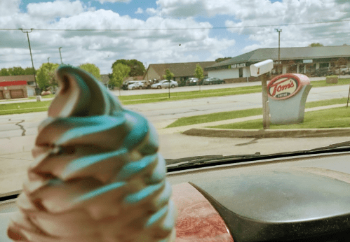 A close-up of a soft-serve ice cream cone with a shop sign in the background on a sunny day.
