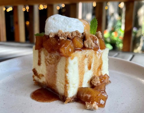 A slice of cheesecake topped with caramel, spiced apples, whipped cream, and a mint leaf on a light plate.