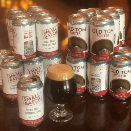 A glass of dark beer in front of stacked cans of Small Batch and Old Tom Porter from Pine River Brewing Co.