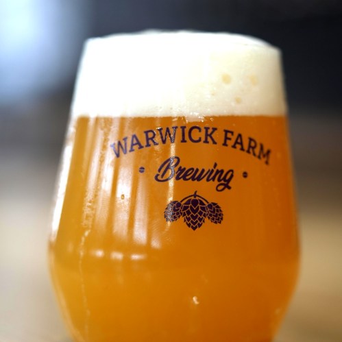 A glass of golden beer with a frothy head, featuring the logo of Warwick Farm Brewing.