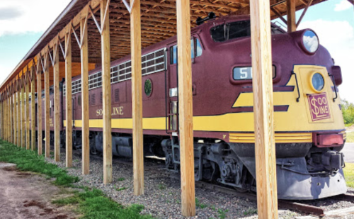 A vintage train displayed under a wooden shelter, featuring maroon and yellow colors.
