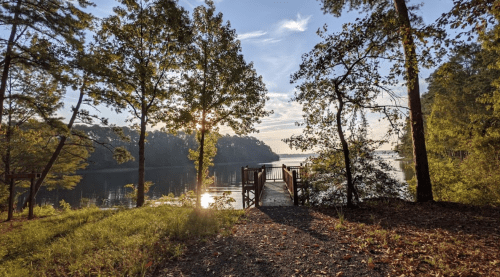 A serene lakeside view at sunset, featuring a wooden dock surrounded by trees and calm water.