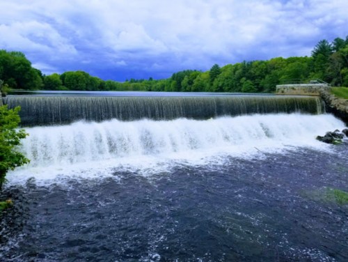 A serene waterfall cascades over a dam, surrounded by lush green trees under a cloudy sky.