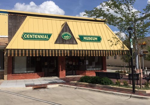 Front view of the Centennial Museum, featuring a yellow triangular roof and green signage, with trees and a sidewalk nearby.