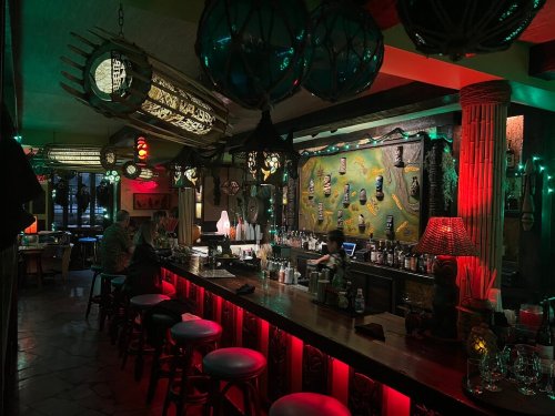 A dimly lit bar with tropical decor, colorful lights, and a bartender preparing drinks.
