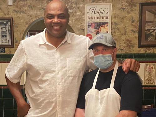 Two men pose together in a restaurant, one wearing a white shirt and the other in a mask and apron.