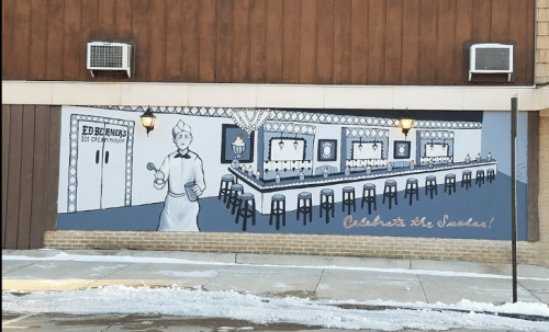 Mural of a vintage ice cream parlor with a server, stools, and the text "Celebrate the Sundae!" on a snowy day.