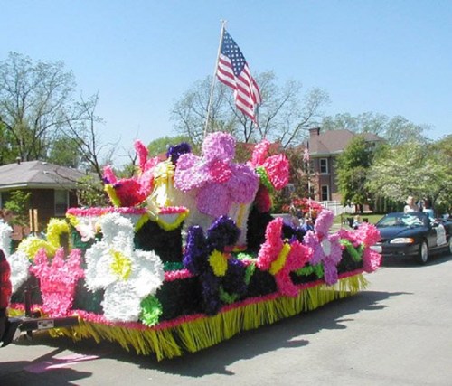 A colorful parade float adorned with large flowers and an American flag, set against a clear blue sky.