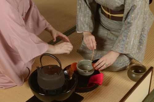 Two people in traditional Japanese attire prepare tea in a serene setting, with a teapot and tea utensils on a tatami mat.
