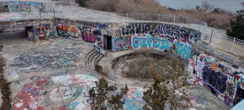 Abandoned circular structure covered in colorful graffiti, surrounded by overgrown vegetation and a view of the water.