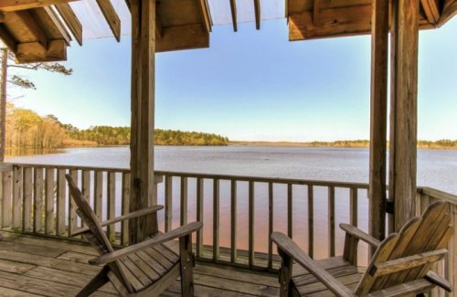 A serene view from a wooden deck overlooking a calm lake surrounded by trees under a clear blue sky.