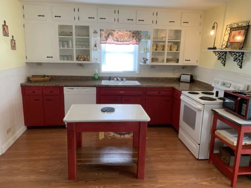 A bright kitchen with red cabinets, a white countertop island, and modern appliances, featuring a window with natural light.