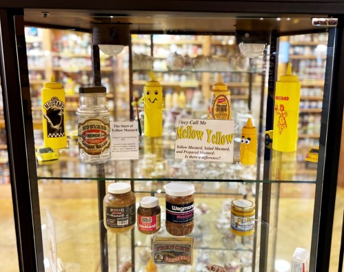 Display case featuring various mustard jars and bottles, including a smiling yellow mustard bottle and a sign about mustard.