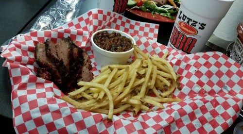 A plate with sliced brisket, fries, and a cup of baked beans, served in a red and white checkered basket.