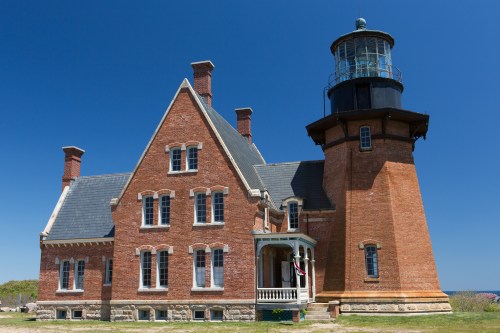 A historic brick lighthouse with a tall tower, surrounded by green grass and a clear blue sky.