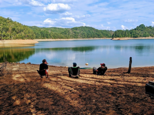 Three people relax by a calm lake surrounded by trees, enjoying the scenic view on a sunny day.