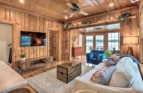 Cozy living room with wooden walls, a TV, comfortable seating, and large windows overlooking a lake.