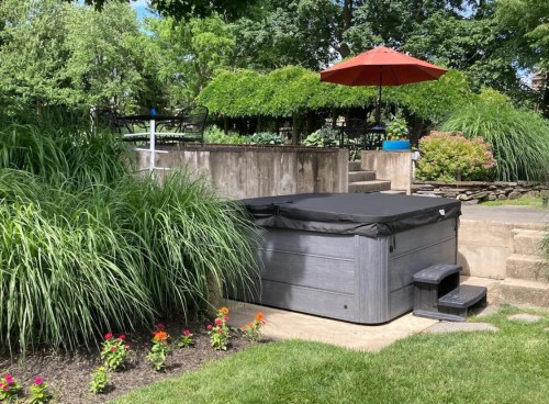 A hot tub with a cover sits beside lush greenery and colorful flowers, shaded by a red umbrella in a serene garden setting.