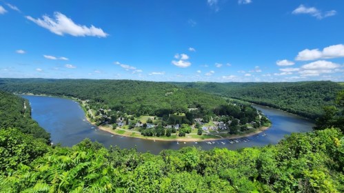 A scenic view of a river winding through lush green hills under a bright blue sky with scattered clouds.