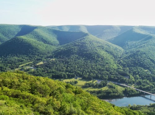 Lush green mountains and valleys under a clear sky, with a river and bridge visible in the foreground.