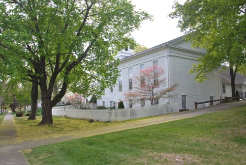A white building surrounded by green trees and a white picket fence, with a path leading up to it.
