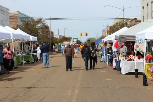 A street market scene with vendors under tents, people walking, and various goods displayed on tables.