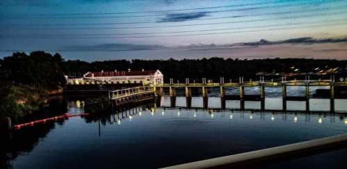 A serene river scene at dusk, featuring a dam, reflections in the water, and power lines in the background.