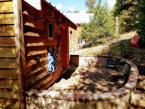 A cozy wooden cabin with a small patio, surrounded by trees and gravel, featuring chairs and towels hanging outside.
