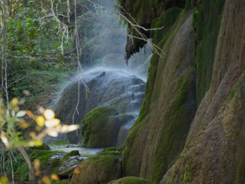 A serene waterfall cascades over moss-covered rocks in a lush, green forest. Mist rises gently in the air.