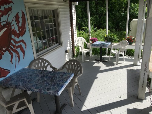 A cozy outdoor dining area with two tables and chairs, surrounded by greenery and a crab mural on the wall.