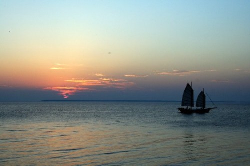 A silhouette of a sailboat on calm waters at sunset, with vibrant colors in the sky reflecting on the surface.