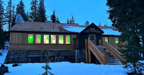 A wooden lodge with colorful lights in the windows, surrounded by snow and evergreen trees at dusk.