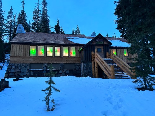 A wooden cabin with colorful lit windows, surrounded by snow and tall trees, under a twilight sky.