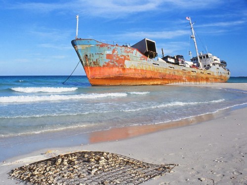 A rusted shipwreck stranded on a sandy beach, with gentle waves lapping at its hull under a clear blue sky.