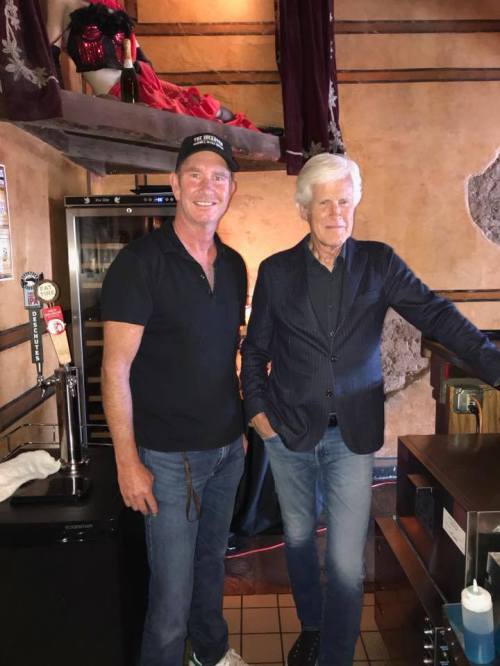 Two men stand together in a restaurant, one in a black shirt and cap, the other in a blazer, smiling at the camera.