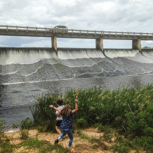 Two children play near a river with a dam and a bridge overhead, surrounded by tall grass and cloudy skies.