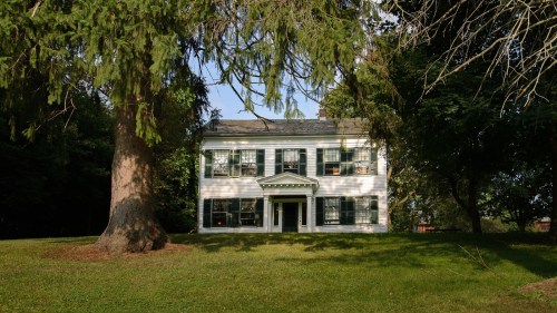 A white two-story house with green shutters, surrounded by trees and a grassy lawn.