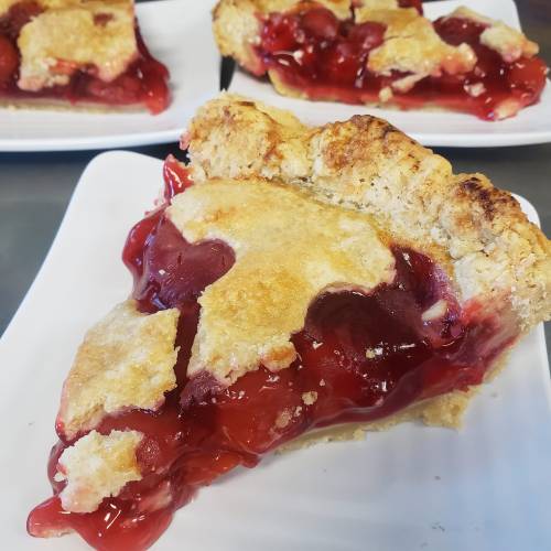 A slice of cherry pie with a golden crust, sitting on a white plate, with more pie slices in the background.