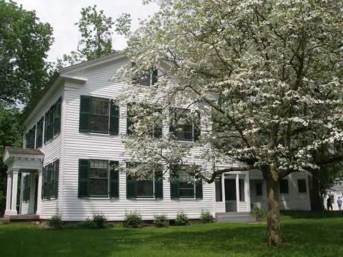 A white two-story house with green shutters, surrounded by blooming trees and a grassy lawn.