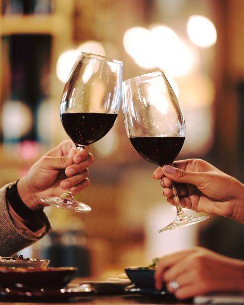 Two hands clink wine glasses filled with red wine, set against a warm, blurred background.