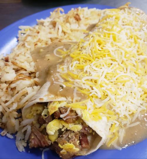 A plate featuring a large breakfast burrito topped with cheese and gravy, alongside crispy hash browns.