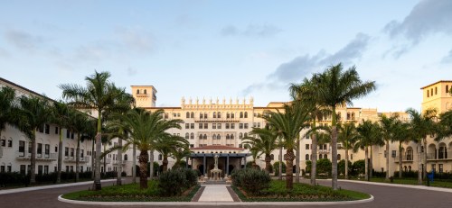 A grand building with palm trees and a fountain in a landscaped courtyard under a clear sky.