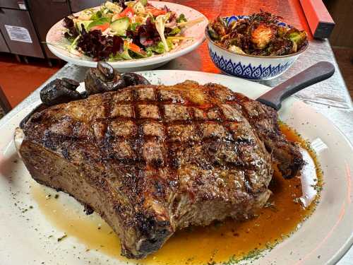 A large, grilled ribeye steak on a plate, accompanied by a fresh salad and a bowl of roasted Brussels sprouts.