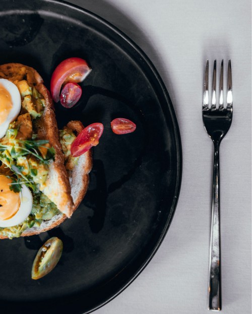 A black plate with avocado toast topped with sliced eggs and cherry tomatoes, alongside a silver fork.