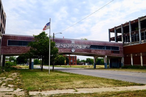 Abandoned Packard plant entrance with a clock, overgrown grass, and a flag in the background under a cloudy sky.